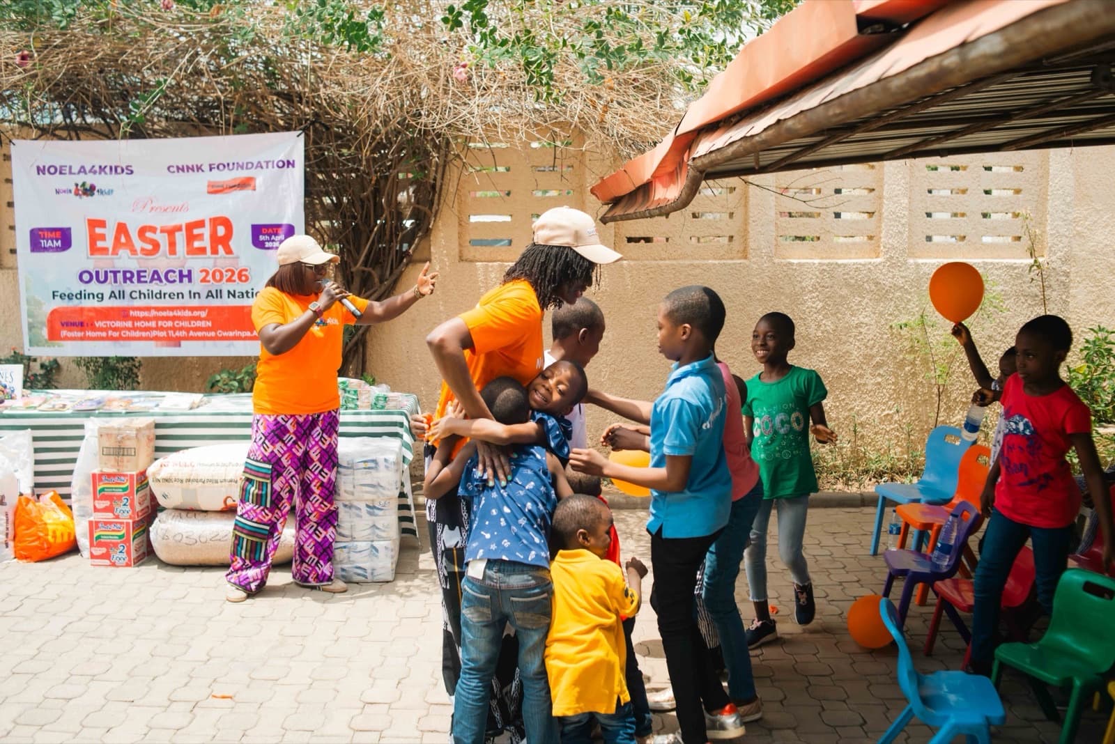 A Noela4kids volunteer welcomes children with a microphone beneath the Easter Outreach 2026 banner